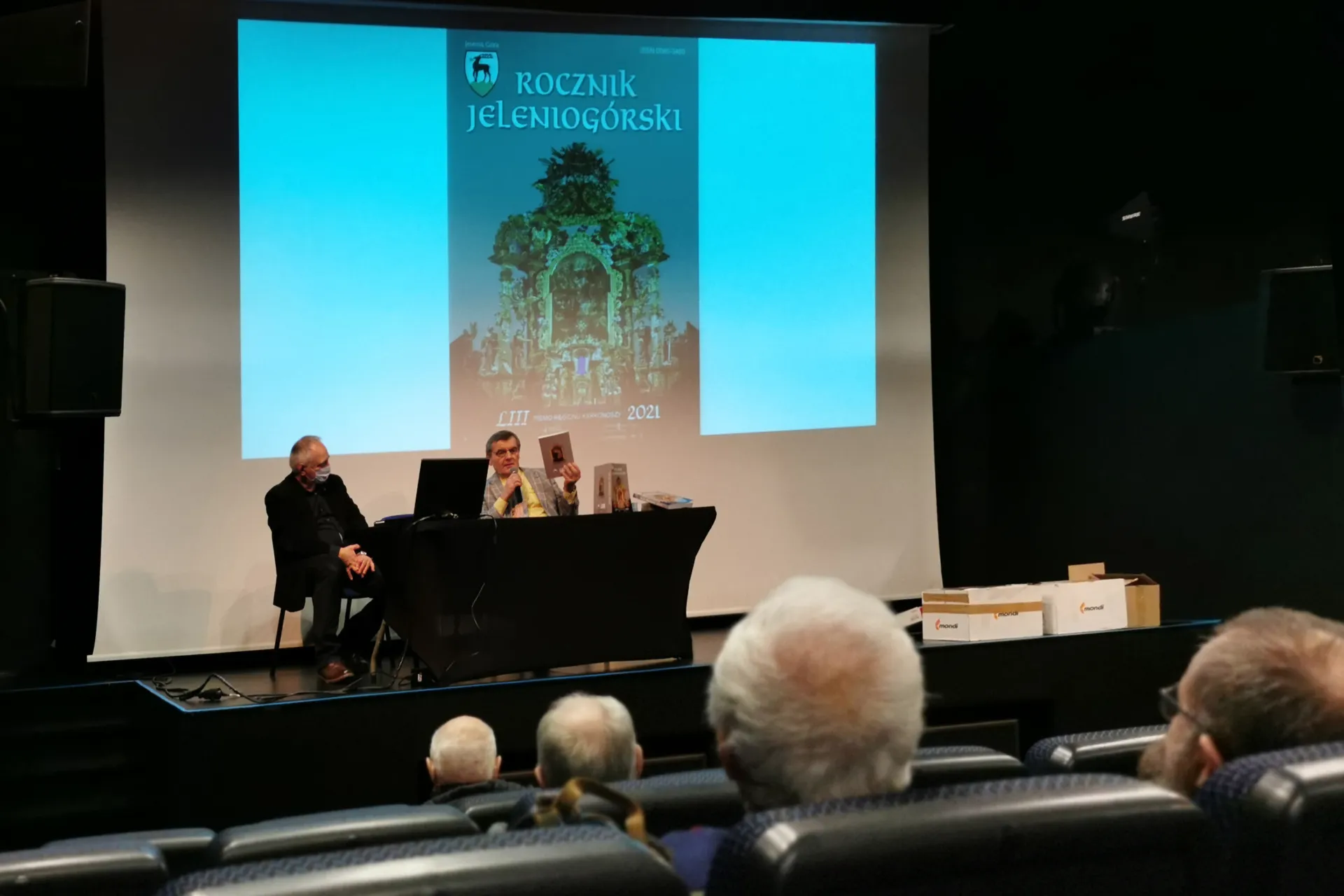 Ivo Łaborewicz i Stanisław Firszt siedzą za stołem konferencyjnym, nad nimi prezentacja z okładką nowego tomu Rocznika Jeleniogórskiego.