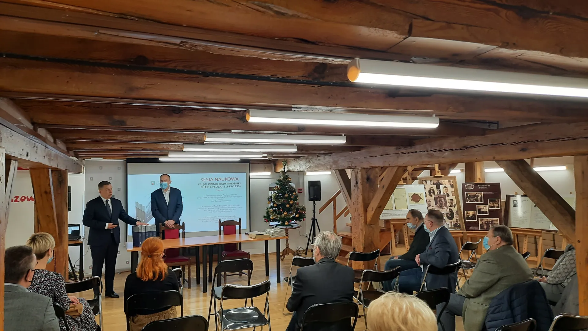 Dyrektor Piekarski i redaktor Gryszpanowicz stoją przy stole konferencyjnym. Przed nimi na krzesłach na Sali siedzi publiczność.
