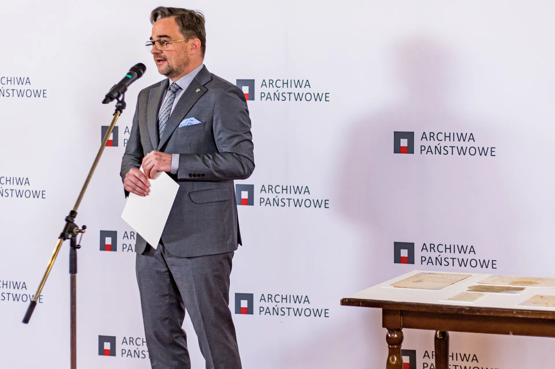 Dyrektor Pietrzyk przed mikrofonem. W tle ścianka z logo Archiwa Państwowe.