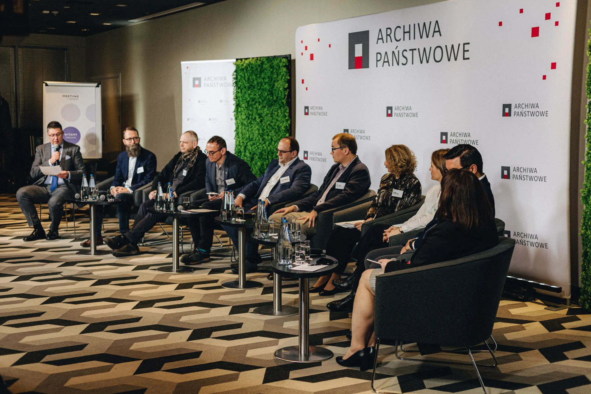 10 osób siedzi na fotelach ustawionych w półokręgu na scenie.