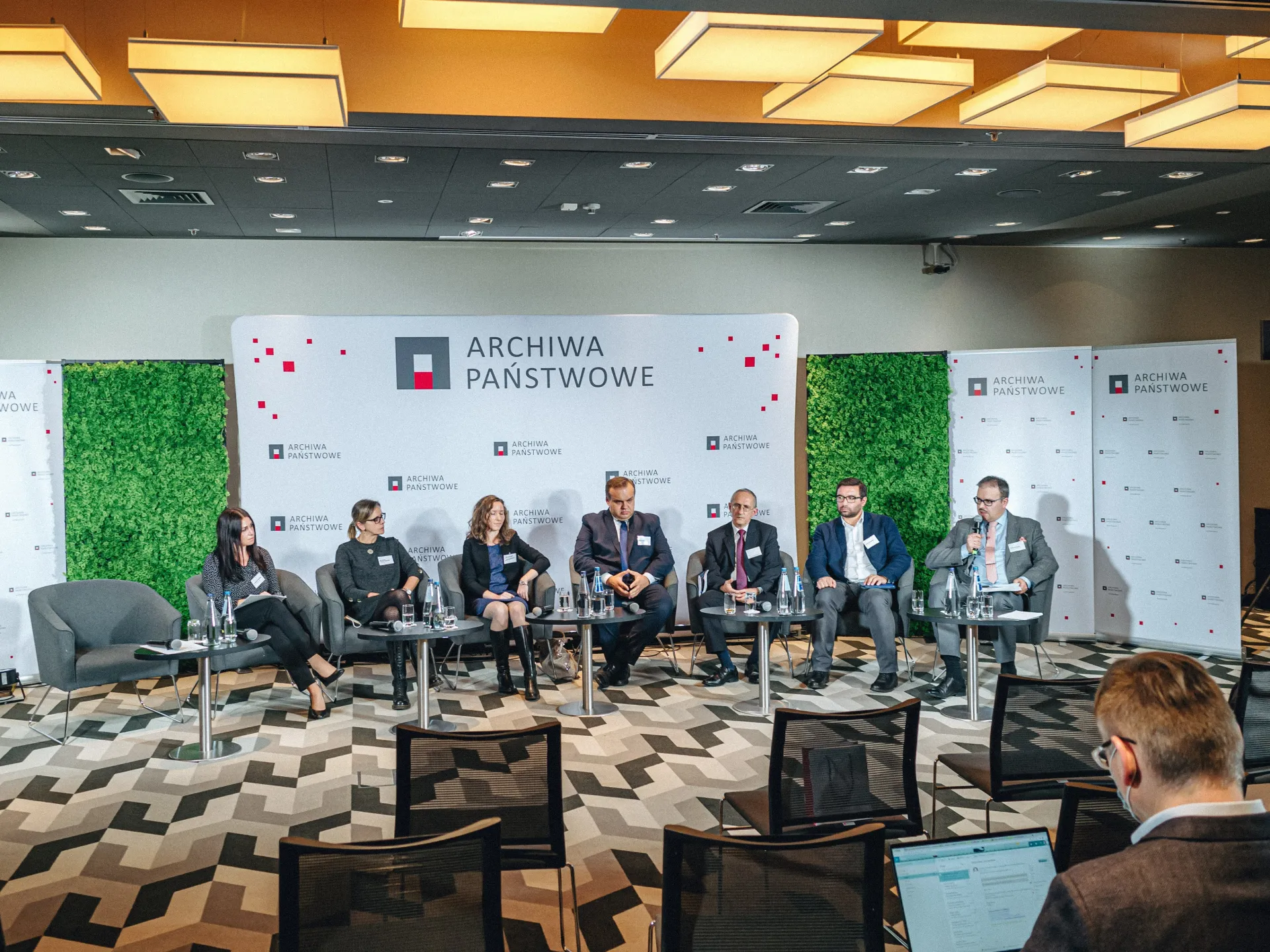 7 osób siedzi na fotelach ustawionych w półokręgu na scenie.
