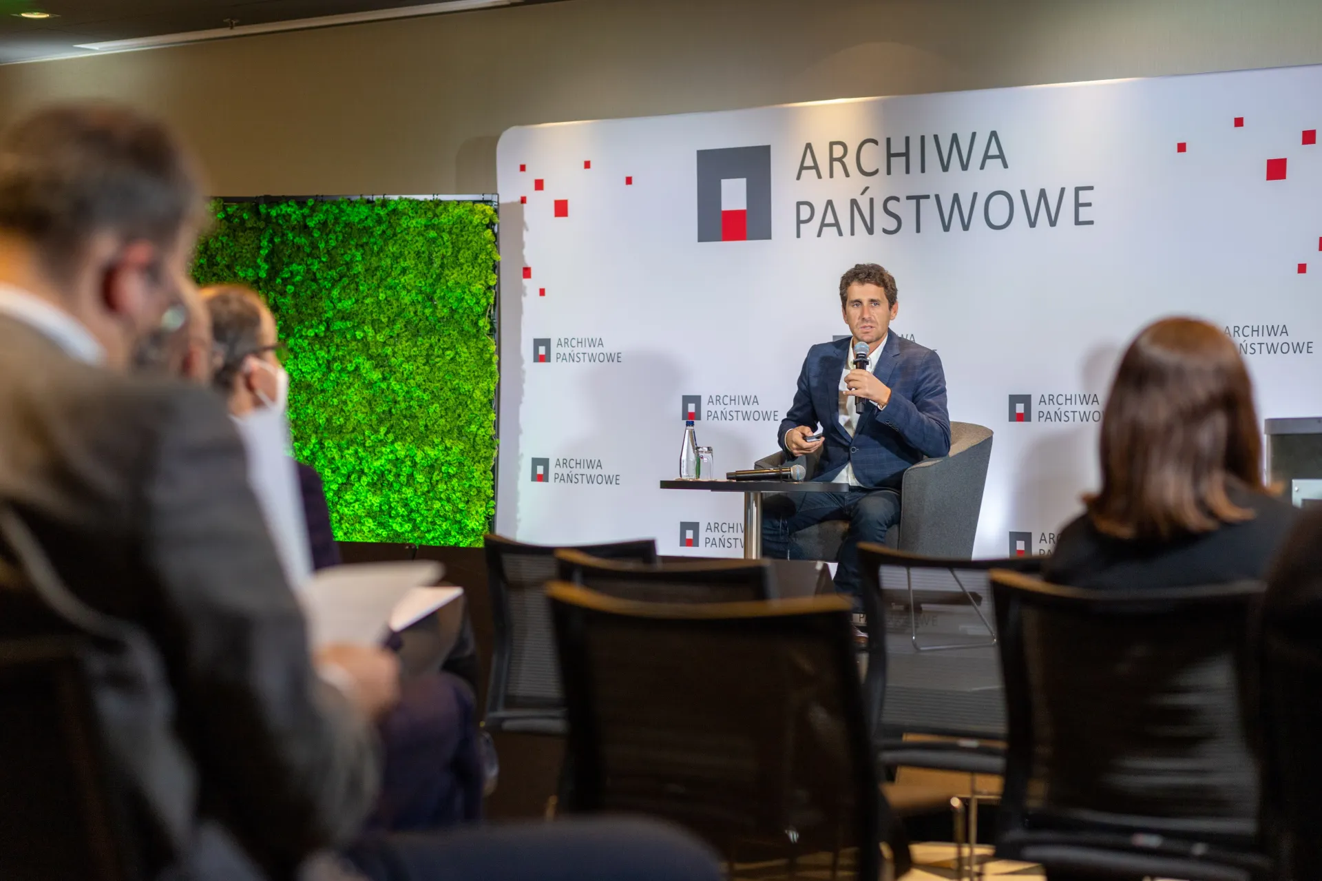 Prelegent Daniel Gomes siedzi na fotelu. W tle ścianka z logo Archiwa Państwowe. Obok przy mównicy stoi prowadzący wydarzenie.