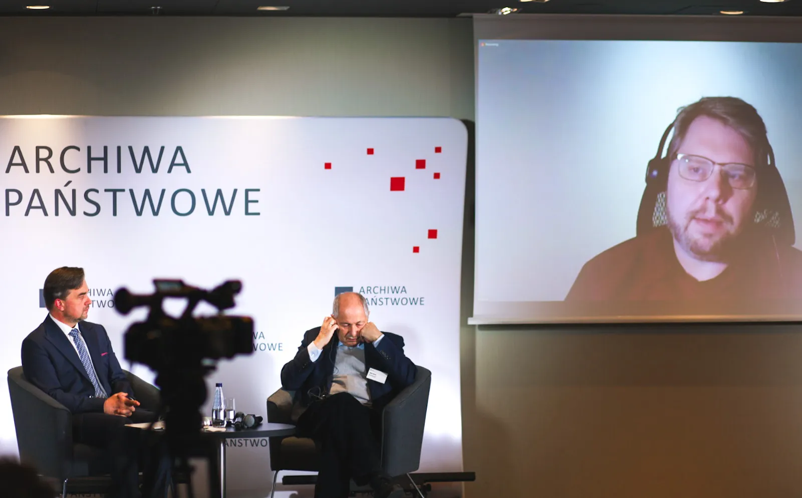 Dyrektor Pietrzyk i Józef Orzeł siedzą na fotelach. W tle ścianka z logo Archiwa Państwowe. Po prawej ekran za zdalnym wystąpieniem Kristinn Sigurðsson.