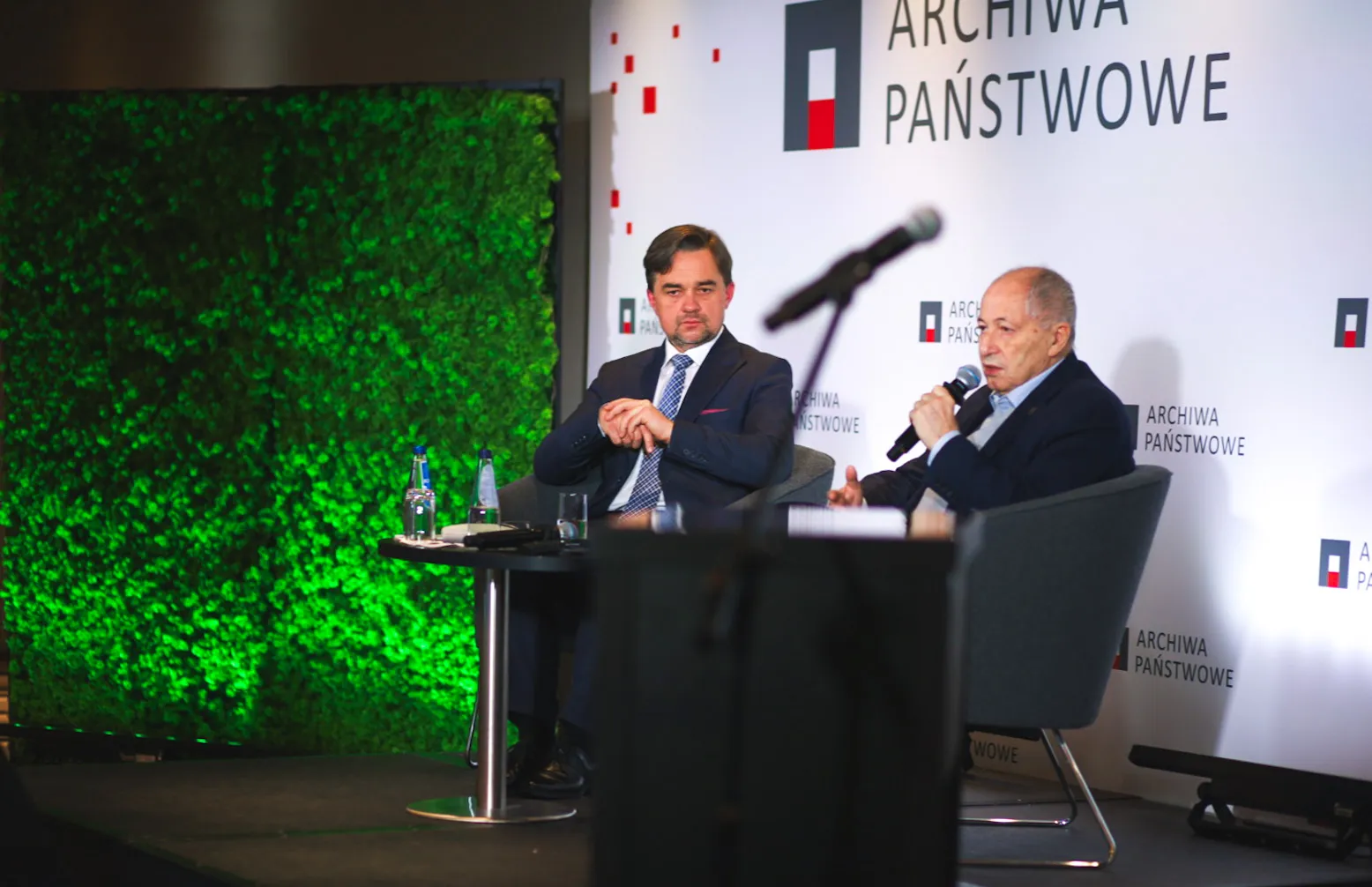 Dyrektor Pietrzyk i Józef Orzeł siedzą na fotelach. Józef Orzeł przemawia do mikrofonu. W tle ścianka z logo Archiwa Państwowe.