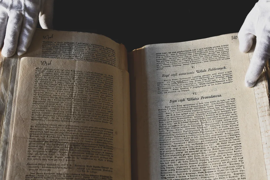Drukowany fragment tekstu Konstytucji 3 Maja trzymany przez osobę w białych, archiwalnych rękawiczkach.