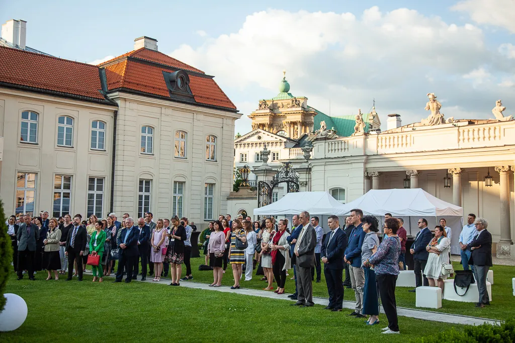 Publiczność stoi na porośniętym trawą dziedzińcu Ministerstwa Kultury, Dziedzictwa Narodowego i Sportu.