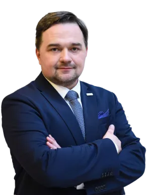 Mężczyzna z krótkimi brązowymi włosami i przystrzyżoną brodą stoi ze skrzyżowanymi ramionami, ubrany w ciemny granatowy garnitur, białą koszulę i krawat w kropki. Ma pewny siebie wyraz twarzy i jest upozowany na prostym białym tle.