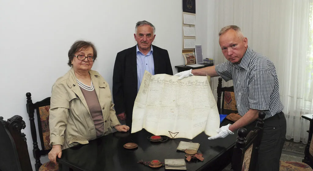 Małgorzata Kaganiec, Zdzisław Jedynak i Janusz Gołaszewski stoją dookoła stołu. Janusz Gołaszewski trzyma w dłoniach rozłożony dokument z pieczęcią. Pozostałe dwa dokumenty leżą złożone na stole. Obok nich pieczęcie.