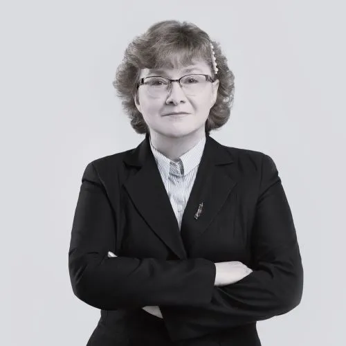 Kobieta w średnim wieku z krótkimi, kręconymi jasnymi włosami, w okularach, ciemnej marynarce i jasnej koszuli z kołnierzykiem, stoi ze skrzyżowanymi ramionami i pewnym siebie wyrazem twarzy na prostym, jasnym tle.