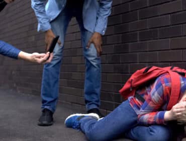Une personne accroupie contre un mur se couvrant la tête tandis qu'une autre personne ramasse un téléphone portable tombé à proximité d'un agresseur potentiel, tous deux impliqués dans la lutte contre la haine en ligne.