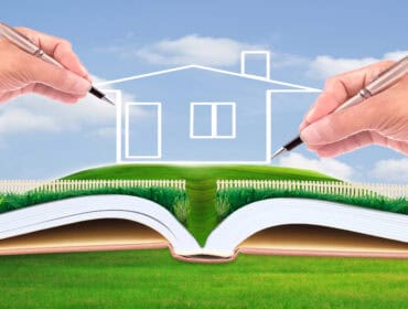 Deux mains dessinant les contours d'une maison au-dessus d'un livre ouvert avec un paysage herbeux, symbolisant le concept de terrain devenu constructible ou de conception architecturale.