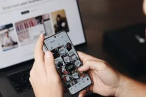 A person holds image-2, a smartphone showing a social media photo grid, with a laptop featuring a blurred screen visible in the background on the desk.