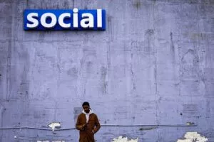 A man in a brown coat stands by a weathered gray wall, looking at his phone. Above him, a large blue sign with "social" in white letters catches the eye. The scene is depicted in the image.