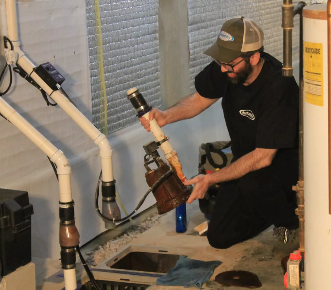 A worker in a black shirt and cap repairs a sump pump in a brightly lit basement. He is holding pump components near white PVC pipes. The mood is focused.