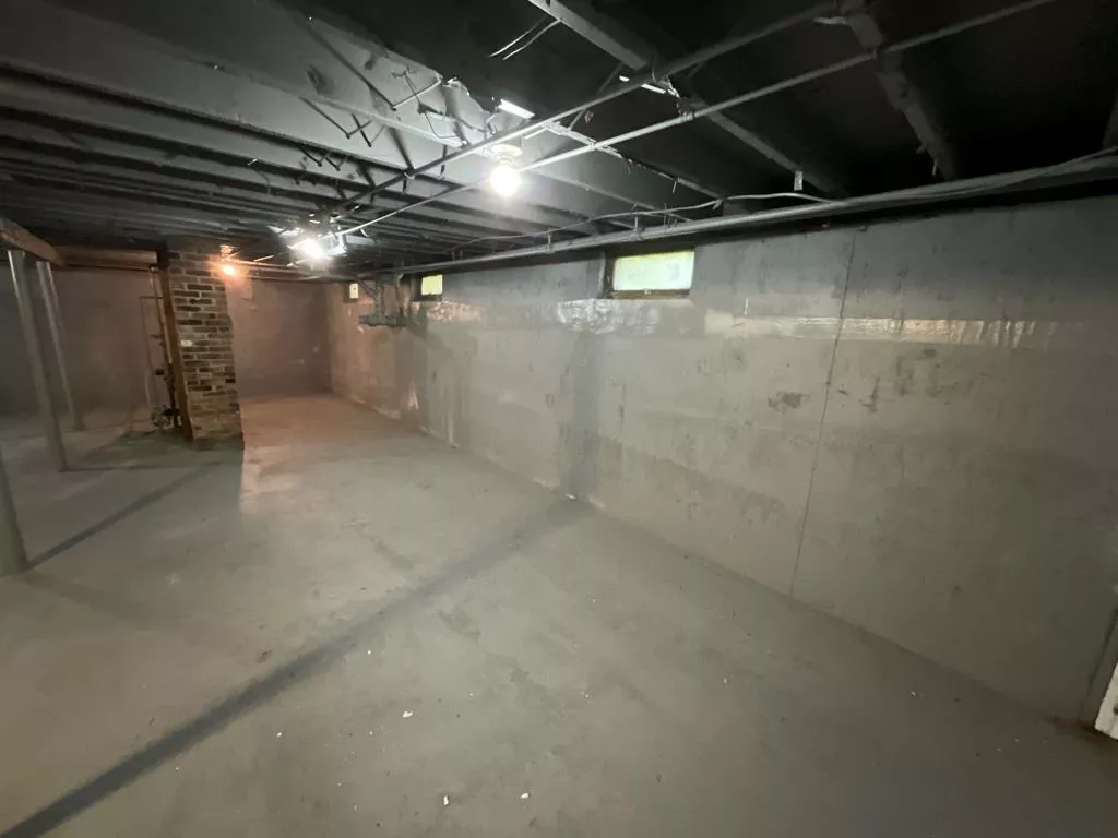 Dimly lit basement with exposed beams and concrete walls. Brick pillar and small windows create a stark, industrial atmosphere.