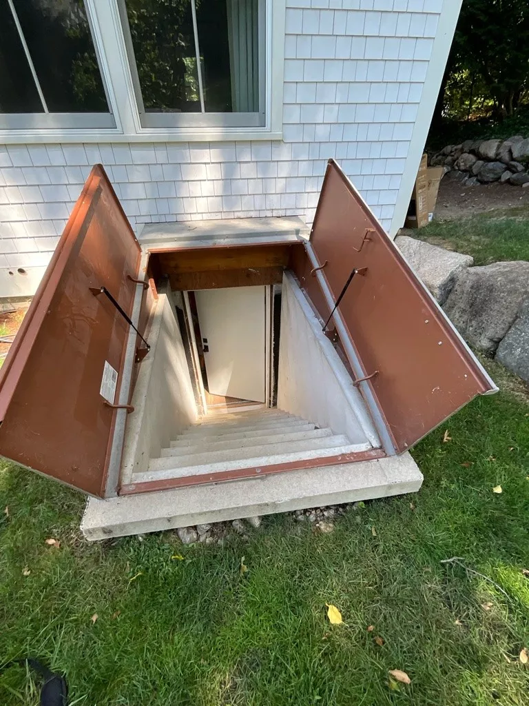 Open cellar doors lead down a set of concrete stairs into a basement. The doors are rusty brown and framed by a grassy lawn, with a house window nearby.