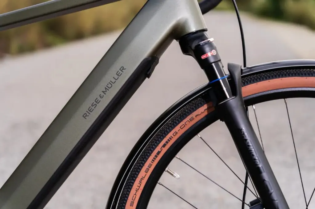 Primer plano de un cuadro de bicicleta Riese & Müller UBN Five con la etiqueta "Riese & Muller", que resalta la ingeniería de precisión de la suspensión delantera y el neumático.