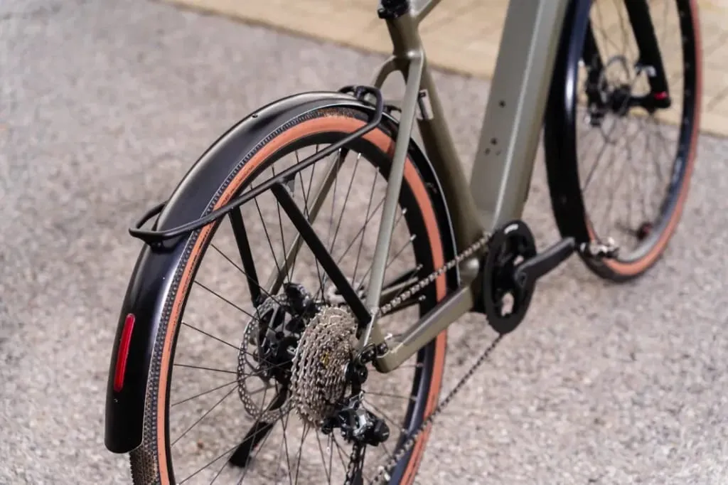 Primer plano de una rueda trasera de bicicleta con un elegante guardabarros urbano, freno de disco y marchas sobre una superficie de grava. Este detalle captura los elementos de diseño innovadores del modelo UBN Five Commute de Riese & Müller.