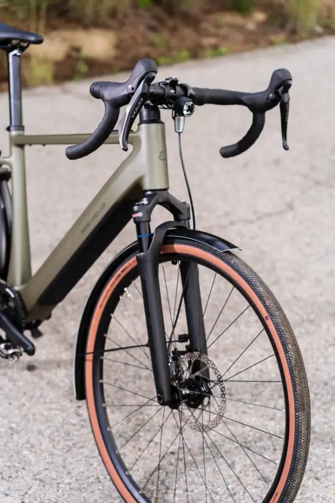 Primer plano de la Riese & Müller UBN Five Commute, que muestra la rueda delantera y el manillar en una carretera asfaltada. El diseño urbano cuenta con un neumático para grava y un freno de disco para un rendimiento mejorado.