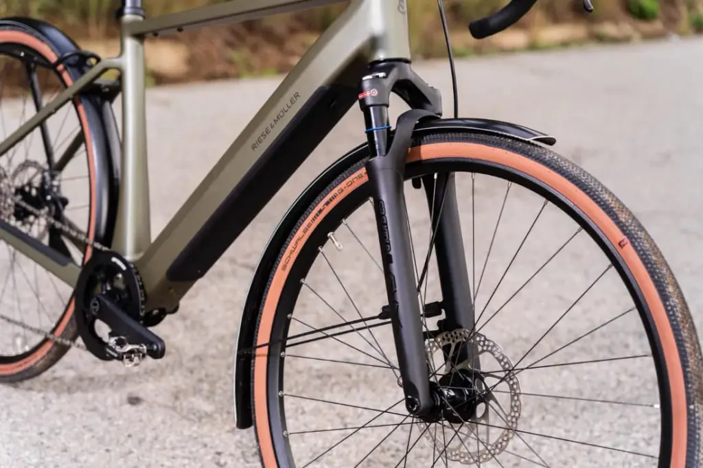 Primer plano de la rueda delantera y la horquilla de una bicicleta Riese & Müller UBN Five Commute con freno de disco, que encarna el espíritu urbano polivalente, estacionada sobre una superficie pavimentada.