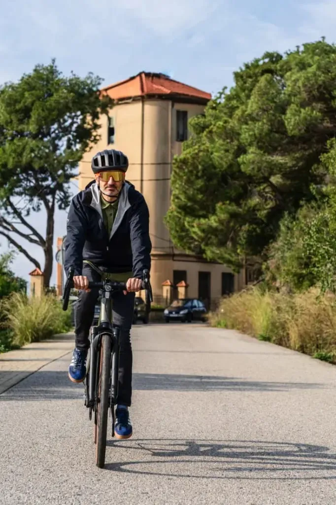 Una persona pasea en una bicicleta Riese & Müller UBN Five Commute por un camino pavimentado, con una torre y árboles que crean un idílico telón de fondo urbano.