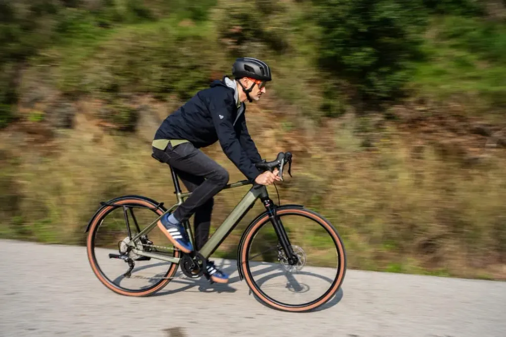 Una persona con casco conduce una bicicleta Riese & Müller UBN Five en un día urbano soleado con una exuberante vegetación de fondo.