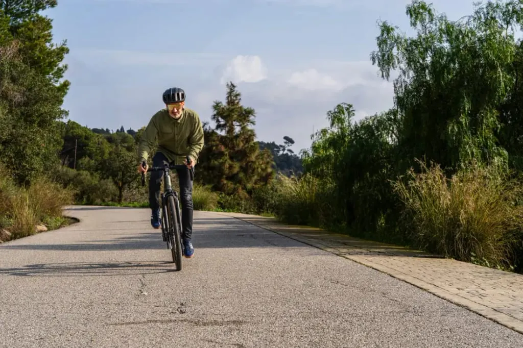 Una persona andando en bicicleta por un sendero soleado, rodeado de árboles y vegetación, se pone un casco y una chaqueta mientras disfruta de su Riese & Müller UBN Five Commute, perfecta para una experiencia urbana polivalente.