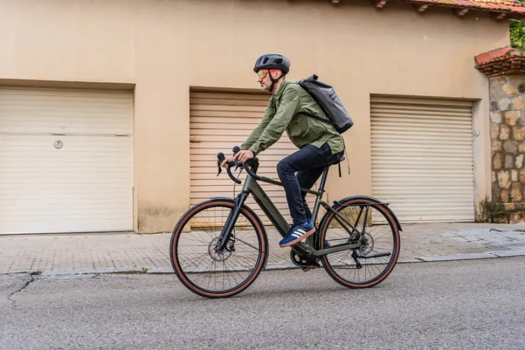 Una persona circula por una calle urbana con una bicicleta Riese & Müller, con casco y mochila, mientras pasa junto a puertas de garaje cerradas.