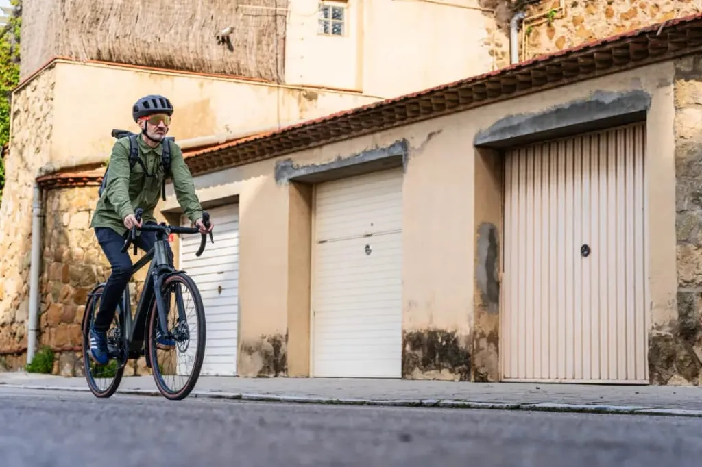 Una persona con casco circula en una bicicleta Riese & Müller UBN Five, mostrando el estilo urbano más polivalente mientras pasa junto a garajes y un edificio de piedra.