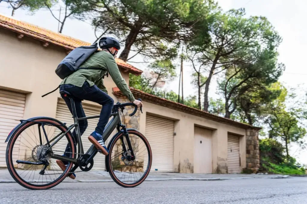 Una persona en una bicicleta Riese & Müller pedalea por una calle urbana cerca de garajes, luciendo un casco y una mochila, rodeada de frondosos árboles.