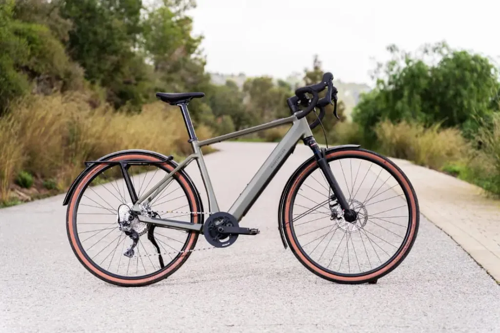 Una elegante bicicleta Riese & Müller UBN Five de color gris, con neumáticos con borde rojo, se desliza por una carretera pintoresca y vacía, flanqueada por una exuberante vegetación.