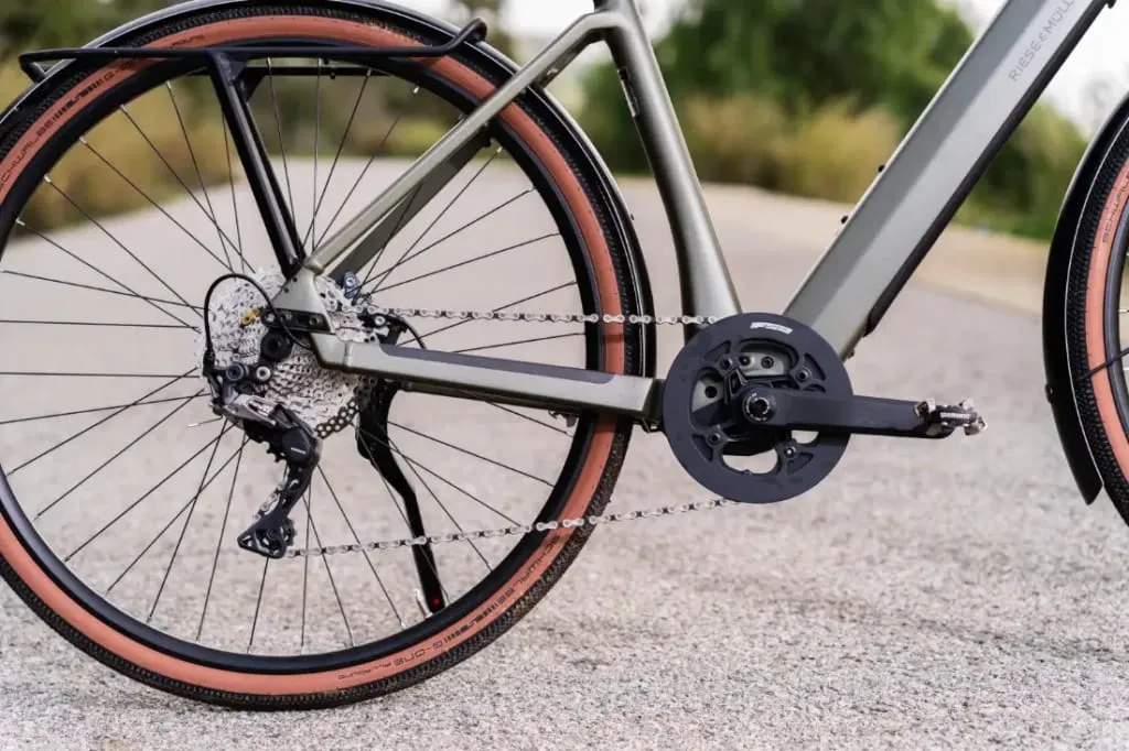 Primer plano de la rueda trasera y la transmisión de la Riese & Müller UBN Five, con neumáticos de paredes color canela, sobre una carretera asfaltada. Esta bicicleta encarna un estilo urbano y, al mismo tiempo, es lo suficientemente polivalente para distintos terrenos.