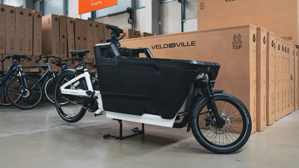 Una bicicleta de carga reposa en el almacén, rodeada de cajas de cartón etiquetadas como "Velo de Ville" junto a otras bicicletas, creando una escena animada que recuerda a los vibrantes mercados de Valencia.