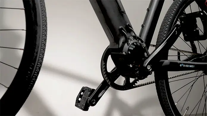 Primer plano de una manivela y un pedal de bicicleta Kosmos Nova7 negra con cadena y rueda trasera visibles sobre un fondo neutro.