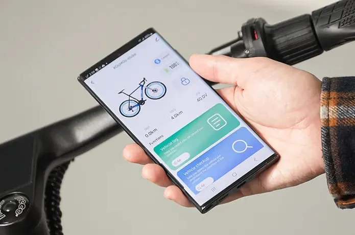 Una persona sostiene un teléfono inteligente kosmos nova7 con una aplicación relacionada con la bicicleta en su pantalla junto al manillar de una bicicleta.