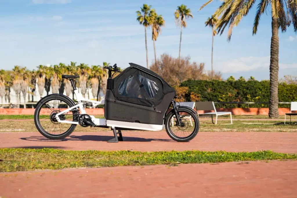 Una elegante Riese & Müller Packster2 70 CT Vario con cubierta negra está estacionada junto a palmeras en un día soleado, ejemplificando la bicicleta de carga eléctrica perfecta para familias urbanas.