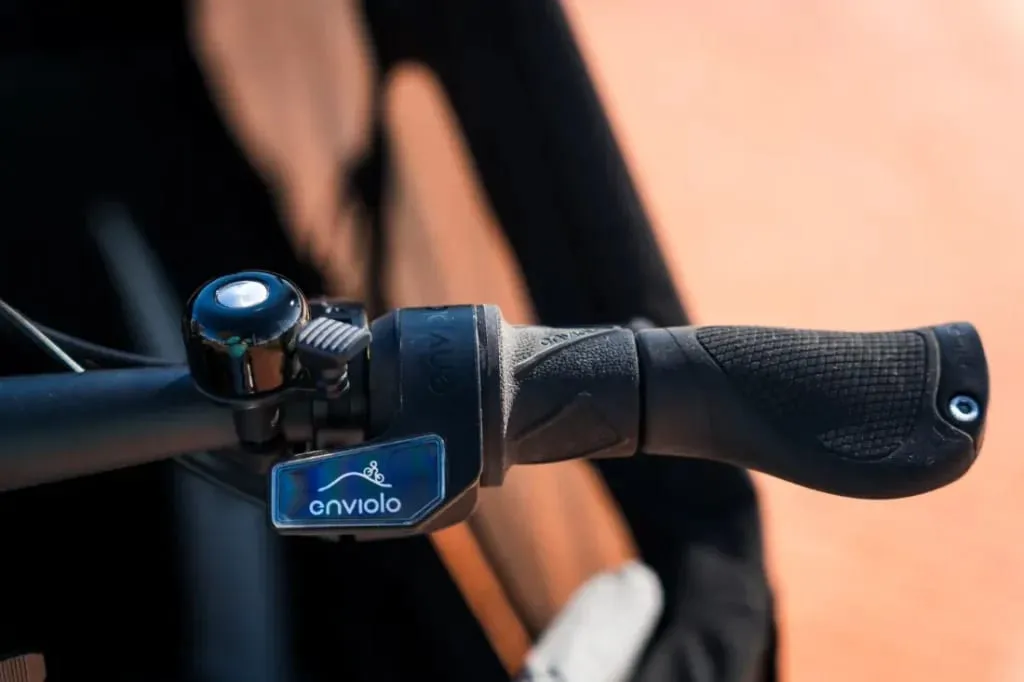 Primer plano de un manillar de bicicleta con un timbre y una palanca de cambios Enviolo sobre un fondo borroso, que muestra el elegante diseño de una bicicleta de carga eléctrica Riese & Müller, perfecta para familias urbanas.