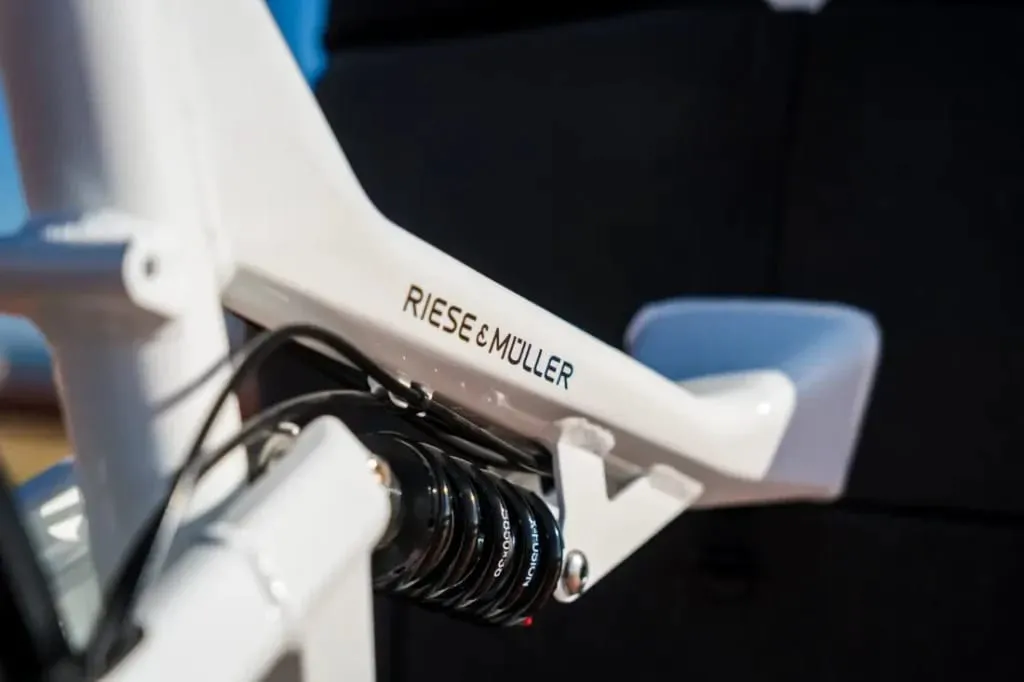 Primer plano del cuadro de una bicicleta de carga eléctrica Riese & Müller blanca que muestra el sistema de suspensión y el logotipo de la marca, perfecto para familias urbanas que buscan soluciones de transporte confiables y elegantes.