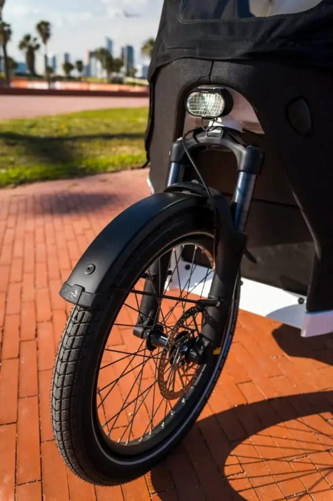 Primer plano de la rueda delantera y el guardabarros de una Riese & Müller Packster2 en un camino de ladrillos, con césped y árboles al fondo, mostrando la bicicleta de carga eléctrica perfecta para familias urbanas.