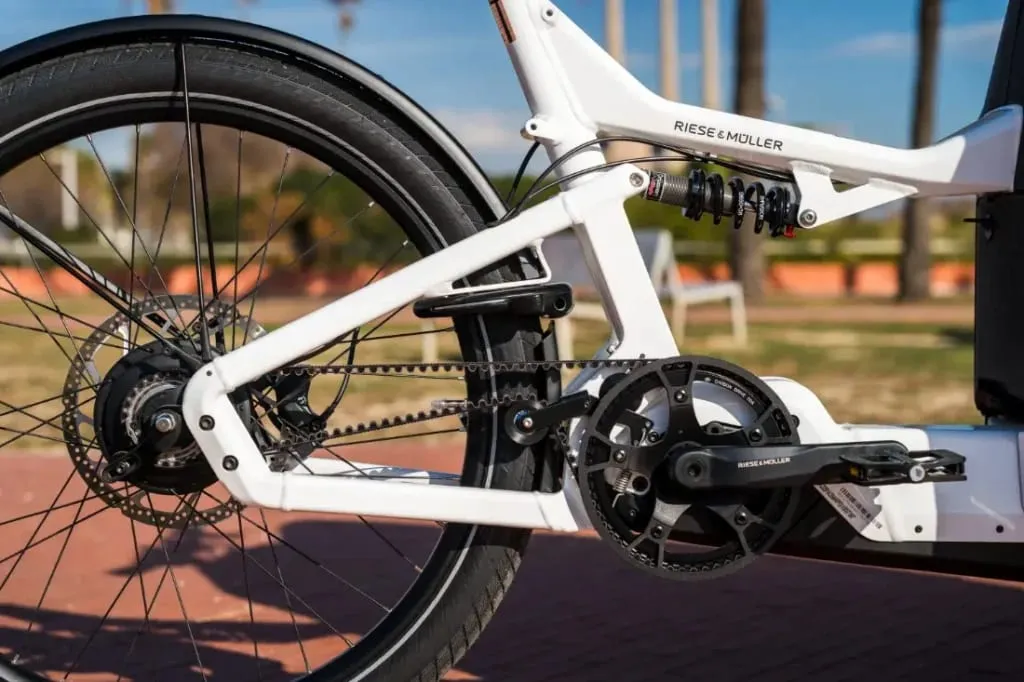 Primer plano del sistema de cadena y suspensión trasera de un Riese & Müller Packster2 70 CT Vario, sobre un fondo de parque, perfecto para familias urbanas que buscan una bicicleta de carga eléctrica eficiente.
