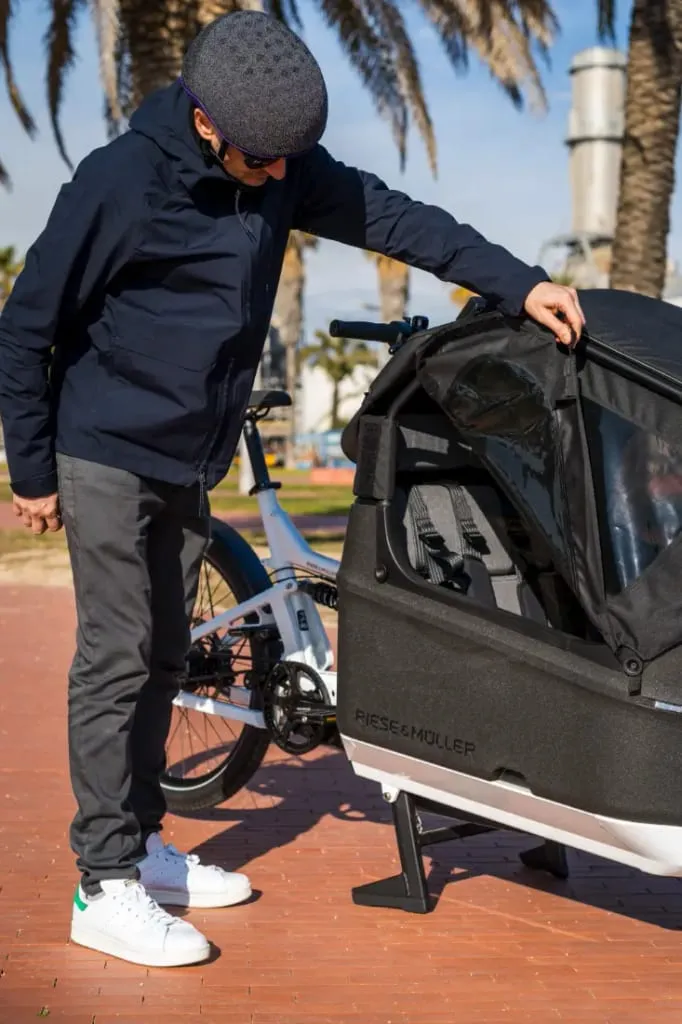 Una persona examina el compartimento del asiento infantil de una bicicleta de carga eléctrica Riese & Müller, perfecta para familias urbanas, bajo palmeras que se balancean en un día soleado.
