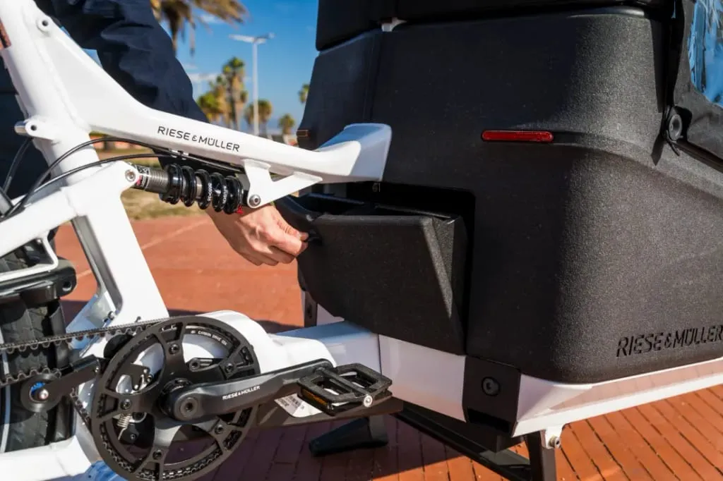 Una persona abre un compartimento en una bicicleta de carga eléctrica Riese & Müller Packster2 70 CT Vario blanca, estacionada en un camino de ladrillos soleado, ideal para familias urbanas que navegan por la vida de la ciudad.