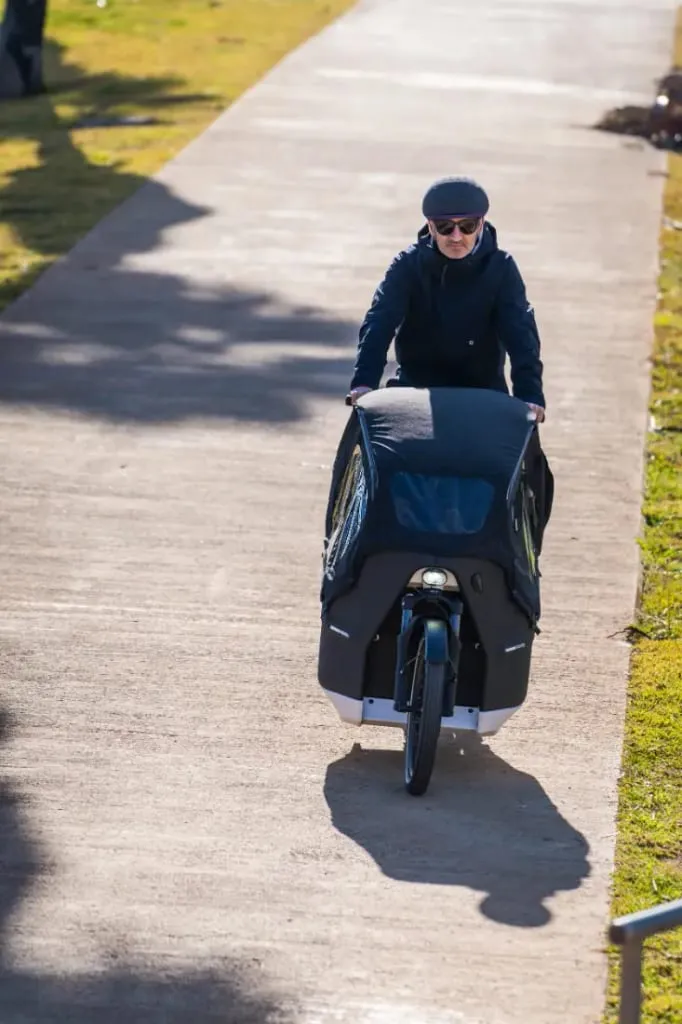 Una persona con gorra y gafas de sol conduce una bicicleta de carga eléctrica Riese & Müller por un sendero urbano soleado.