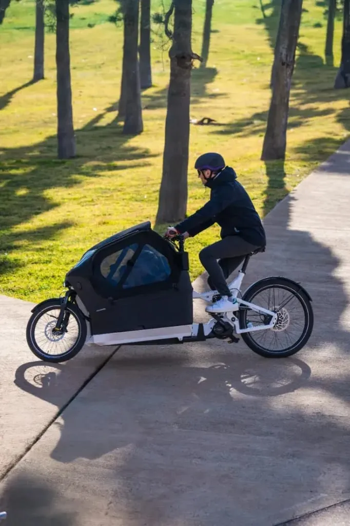 Una persona viaja en una Riese & Müller Packster2 70 CT Vario, su bicicleta de carga eléctrica se desliza suavemente por el sendero soleado del parque, integrándose perfectamente al ritmo de las familias urbanas que disfrutan el día.