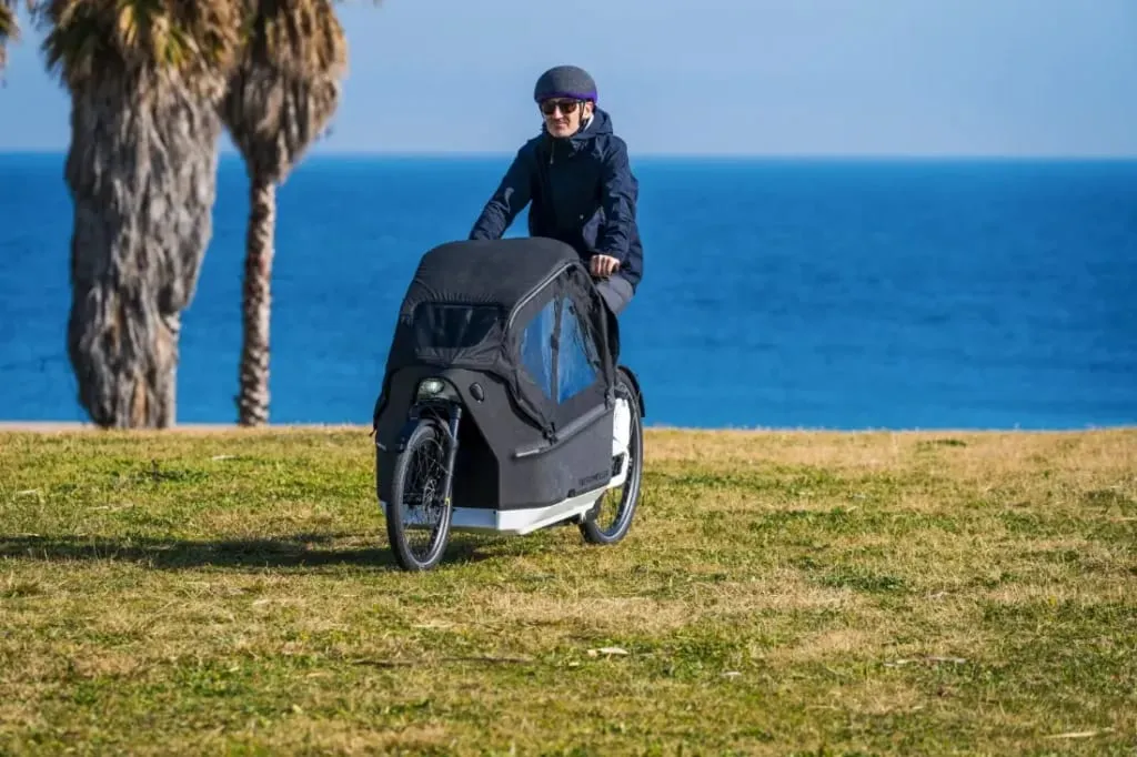 Una persona conduce una bicicleta de carga eléctrica Riese & Müller Packster2 70 CT Vario, con una cesta delantera cubierta, por una zona verde cerca del océano. Perfecta para familias urbanas, esta bicicleta combina estilo y utilidad con el impresionante telón de fondo costero.
