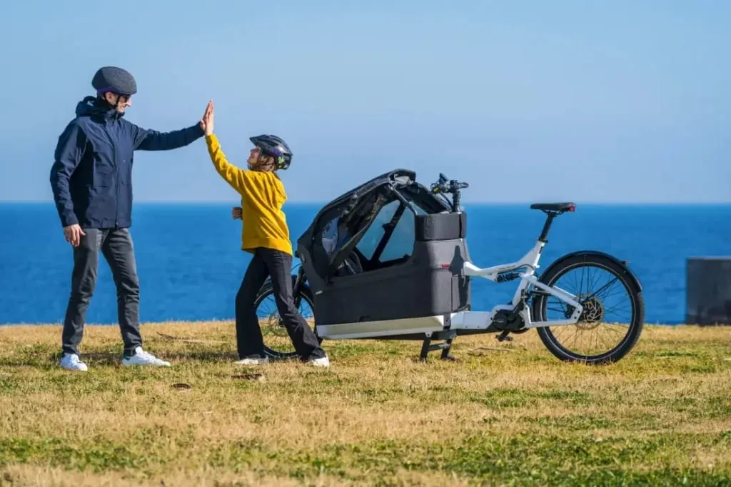 Un niño y un adulto chocan las manos cerca de una bicicleta de carga eléctrica Riese & Müller Packster2 70 CT Vario en un campo de hierba junto al mar, lo que refleja la alegría cotidiana de las familias urbanas.