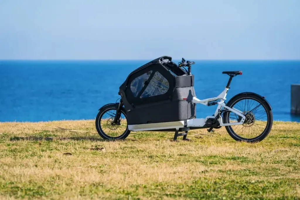 Una bicicleta de carga eléctrica Riese & Müller con una elegante cubierta negra reposa elegantemente sobre el césped junto al mar; su presencia se mezcla armoniosamente con el cielo azul claro: una compañera perfecta para familias urbanas que exploran aventuras costeras.