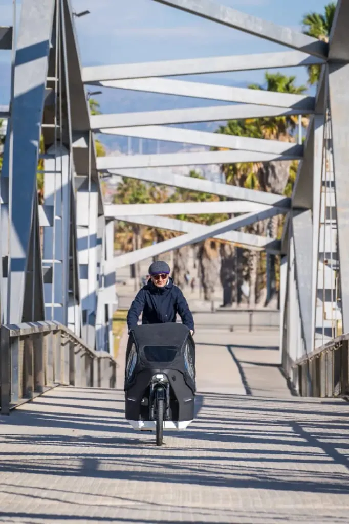 Una persona conduce una bicicleta de carga eléctrica Riese & Müller Packster2 70 CT Vario por un puente, con gafas de sol y una gorra. Al fondo se vislumbra unas palmeras y unas montañas que crean la escena perfecta para familias urbanas.