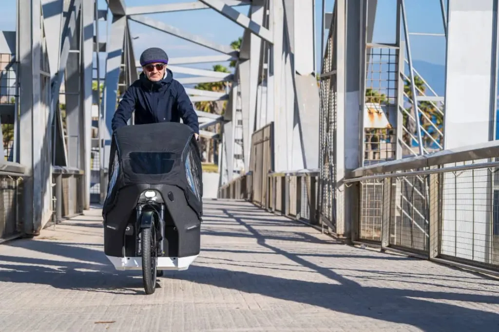Una persona con ropa de invierno viaja en una bicicleta de carga eléctrica Riese & Müller Packster2 a través de un puente de metal, integrándose perfectamente al paisaje de familias urbanas.