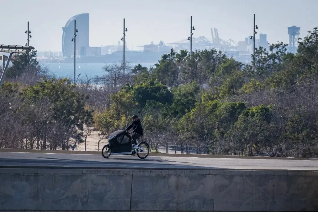 Una persona en una bicicleta de carga eléctrica Packster 70 CT Vario se desliza por el camino, mezclándose a la perfección con el paisaje urbano y el océano en el fondo, sirviendo como una conexión armoniosa entre las familias urbanas y la naturaleza.